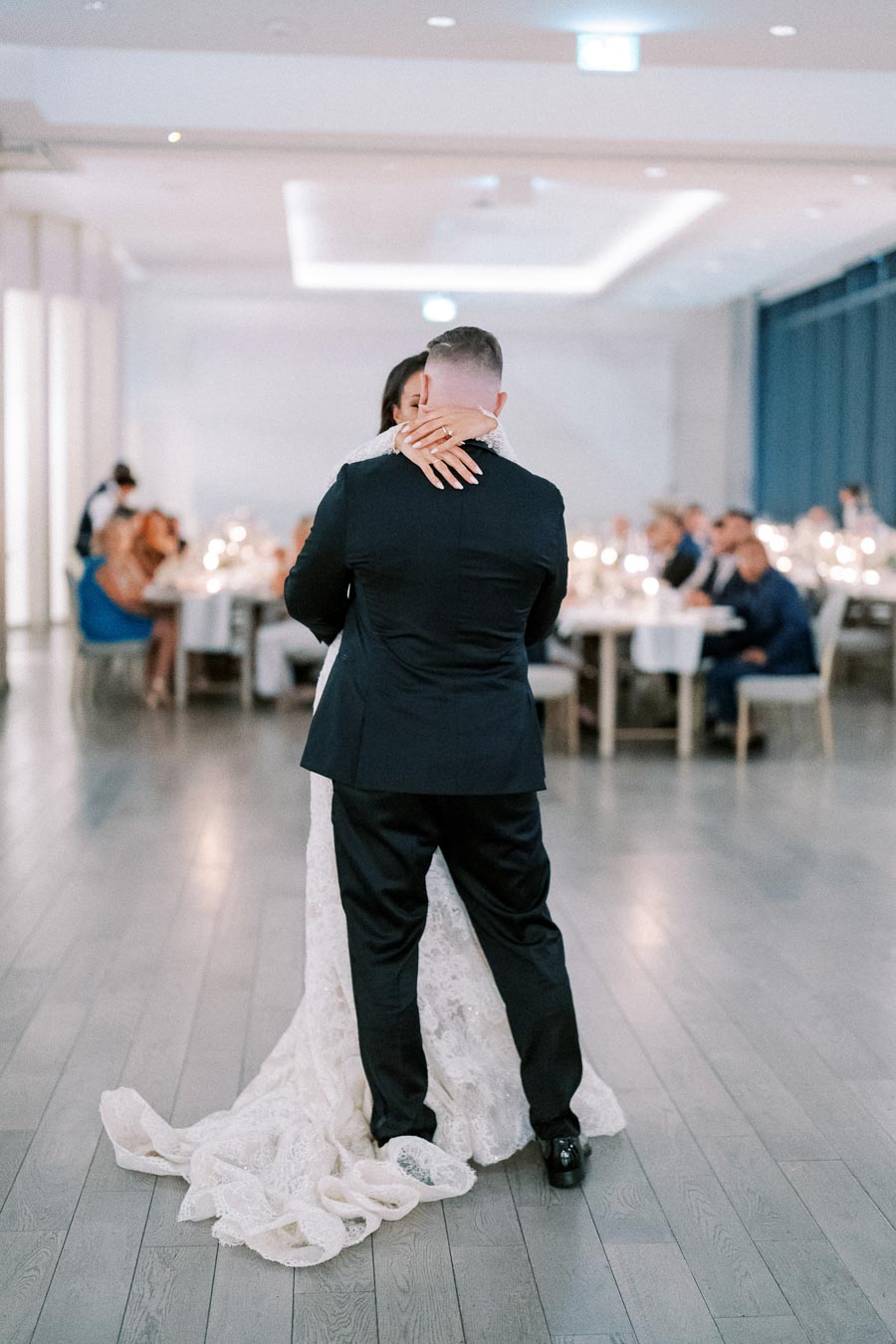 A couple sharing their first dance at a wedding reception in an elegant, softly lit venue, with guests seated at tables in the background.