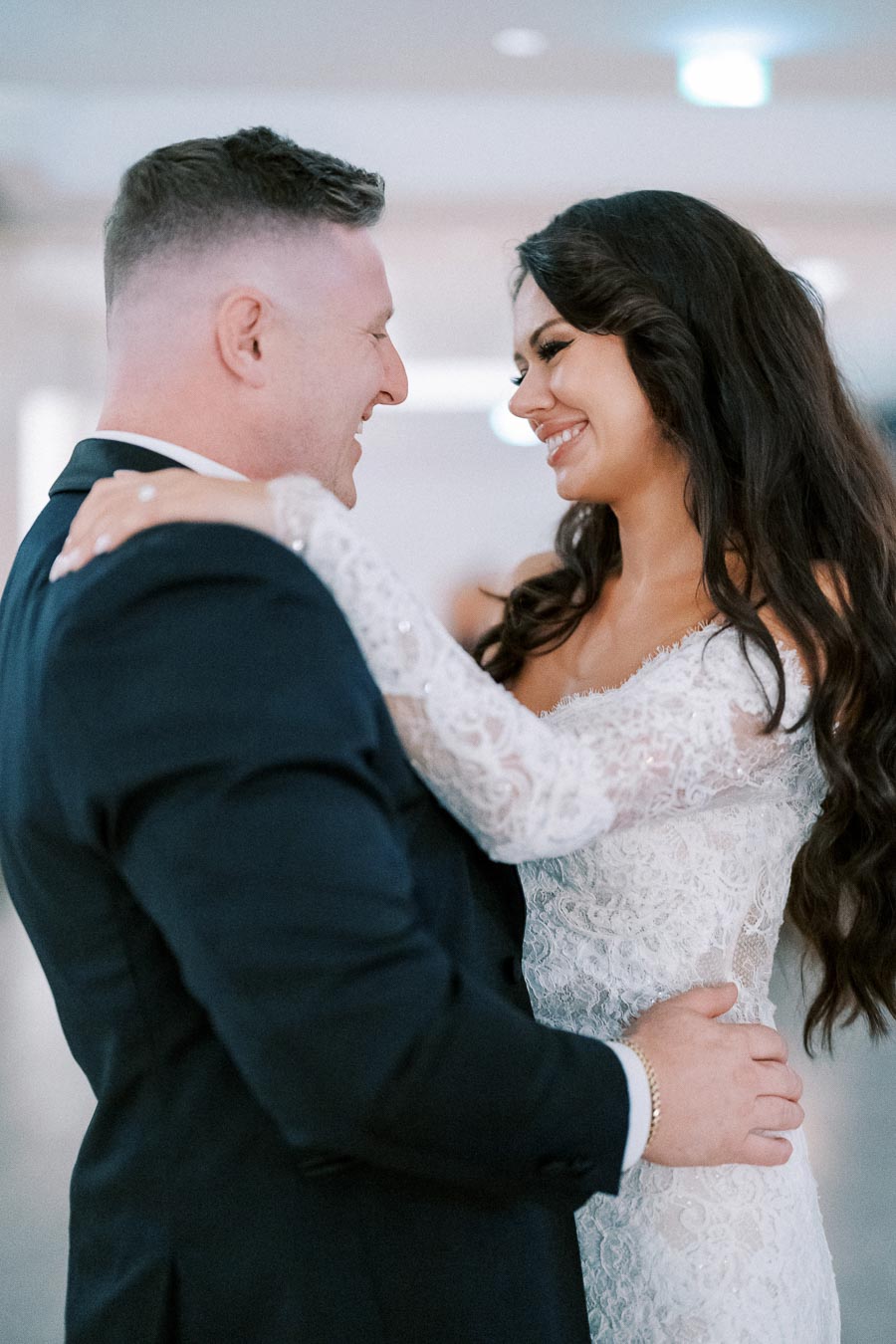 Smiling bride and groom dancing at their wedding reception, wearing elegant attire and embracing closely.