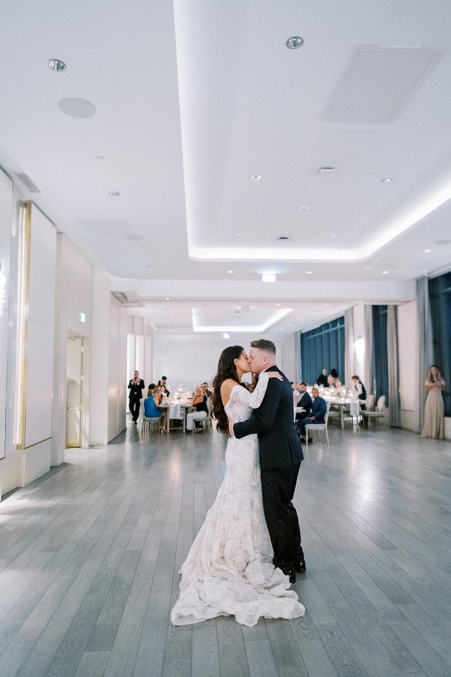 Wedding reception first dance: bride in lace gown and groom in black suit embracing in a modern, elegant venue with guests seated in the background.