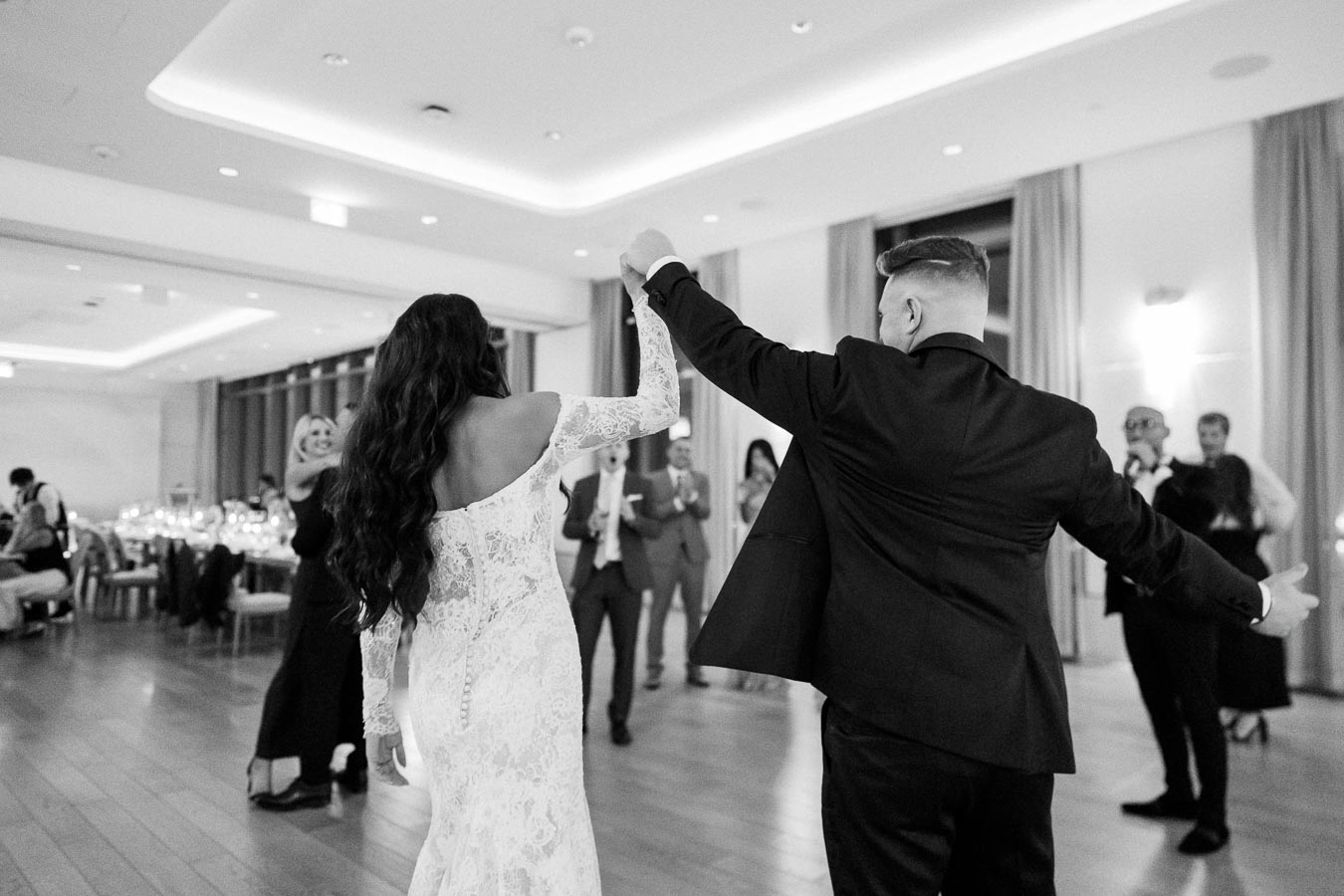 A bride in a lace wedding dress and a groom in a formal suit dance together in a beautifully lit reception hall, surrounded by smiling guests.