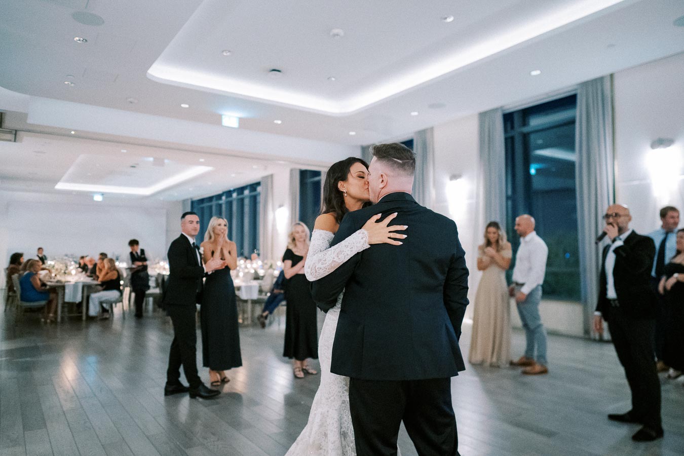 A bride and groom share a romantic first dance at an elegant indoor wedding reception filled with guests in formal attire.