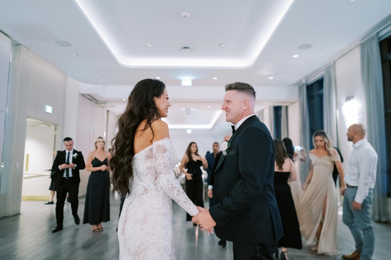 A bride and groom holding hands and smiling at each other during their first dance at a wedding reception, surrounded by guests in an elegantly decorated venue.