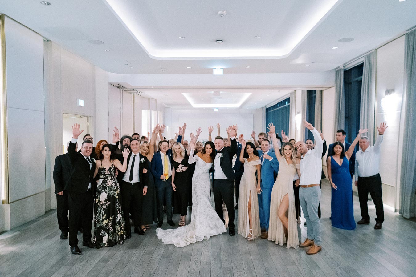 A joyful group photo at a wedding reception, featuring a diverse mix of elegantly dressed guests celebrating with raised hands in a modern and well-lit venue.