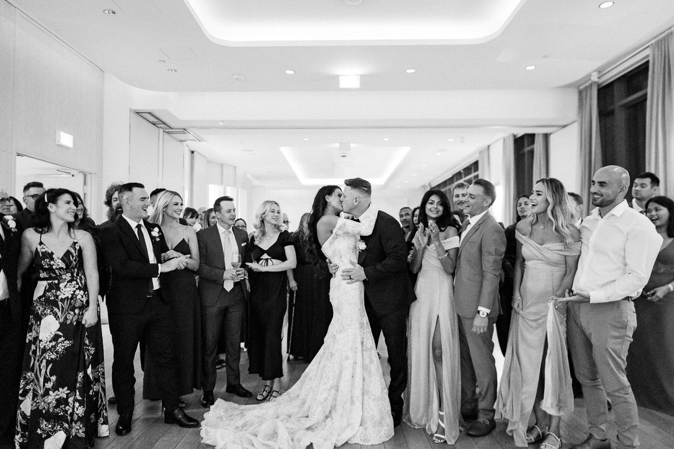 A bride and groom share a kiss in the center of a joyful wedding reception with guests applauding around them. The black and white photo captures the celebration atmosphere and elegant attire of the attendees.
