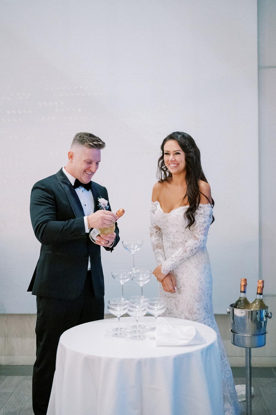 A bride and groom in formal attire smiling while preparing to pour champagne into a tower of glasses at their wedding reception.