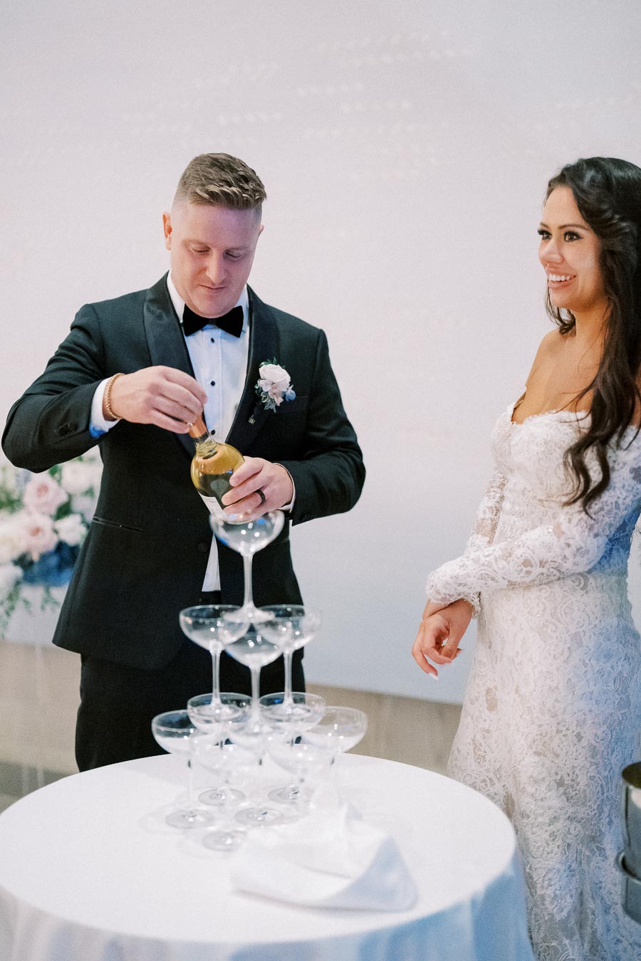 A groom in a black tuxedo pours champagne into a tiered glass tower, while the bride in a white lace wedding dress smiles beside him. The elegant setup is part of a wedding celebration, with flower arrangements visible in the background.