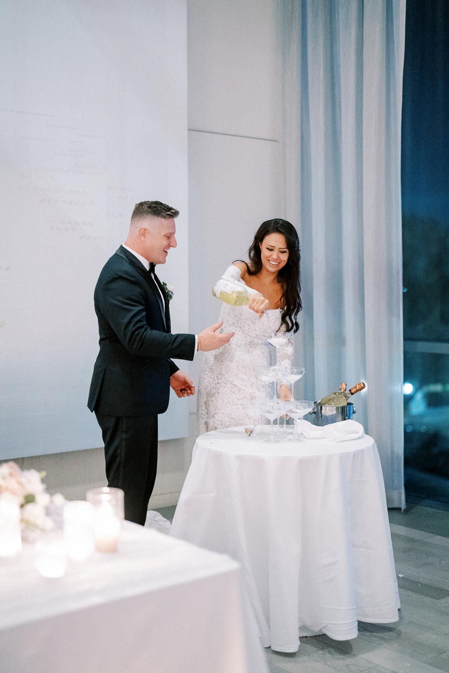 A couple celebrating their wedding by pouring champagne into a stacked glass tower at a reception.
