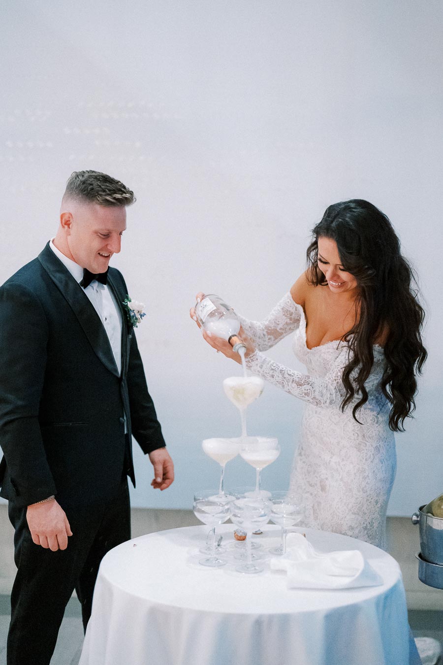 A bride in a white lace dress and a groom in a black tuxedo celebrate by pouring champagne into a cascading tower of glasses on their wedding day, creating a joyful and elegant moment.