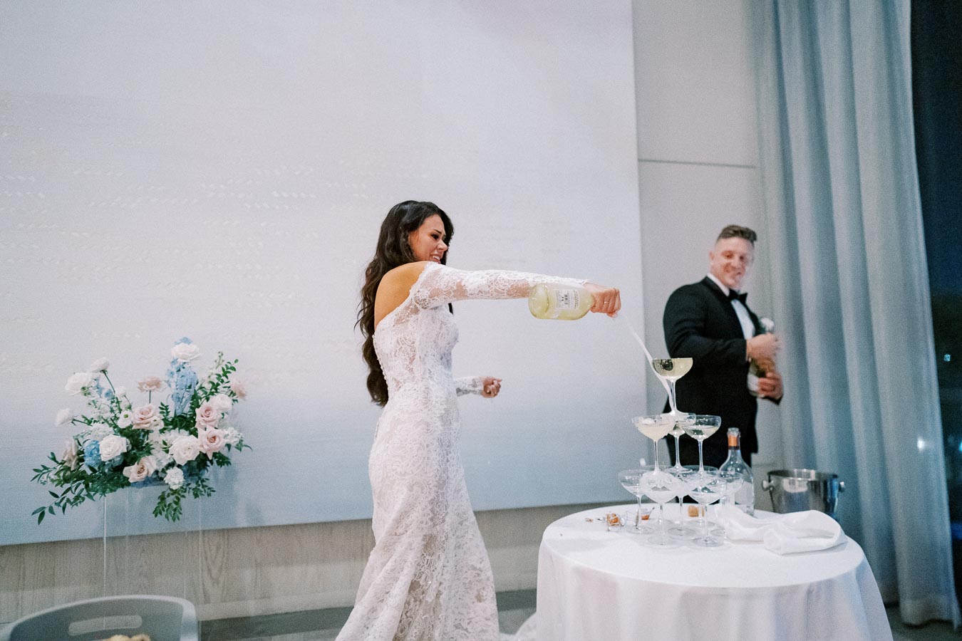 A bride in a lace wedding dress pours champagne into a pyramid of glasses, smiling, with a groom in a tuxedo standing nearby, against a backdrop of elegant decor with pastel flowers.