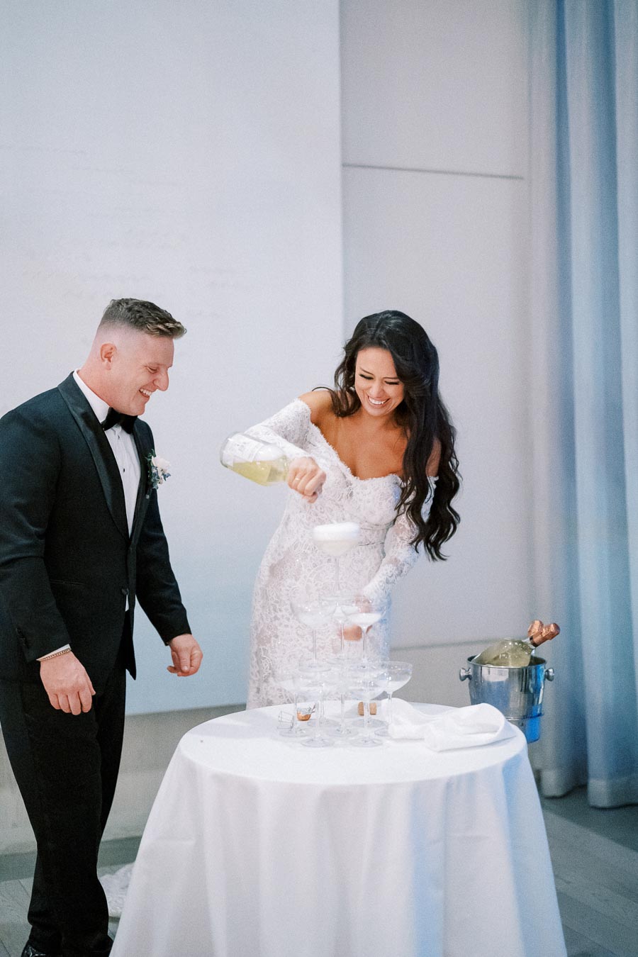 A bride in a lace wedding dress joyfully pours champagne into a tower of glasses, while the groom in a black tuxedo watches and smiles.