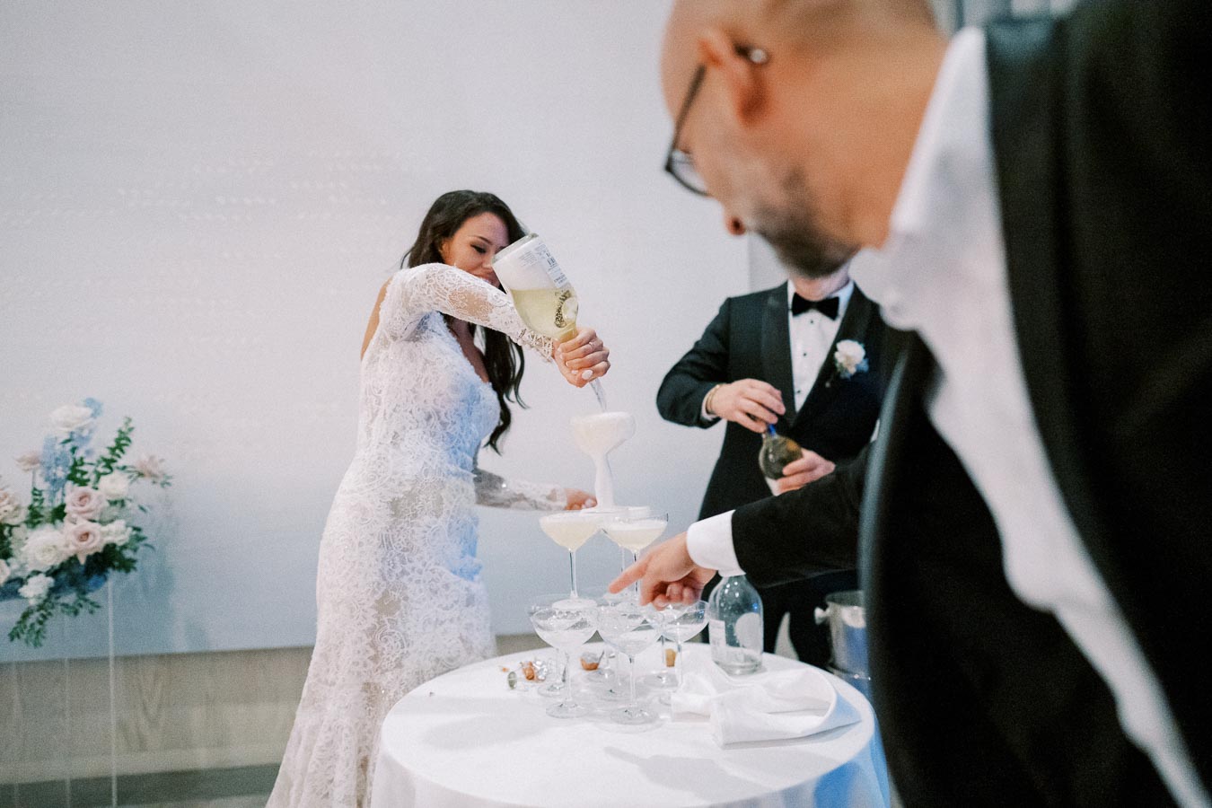 Elegant bride in a lace gown pouring champagne into a cascading tower of glasses during a wedding celebration, with two men in tuxedos assisting.