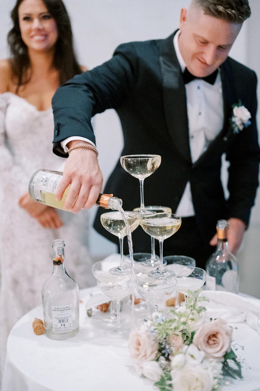 A groom in a tuxedo pours champagne into a tower of glasses, with a bride in a white wedding dress smiling in the background. A bouquet of flowers rests on the table beside champagne bottles and corks.
