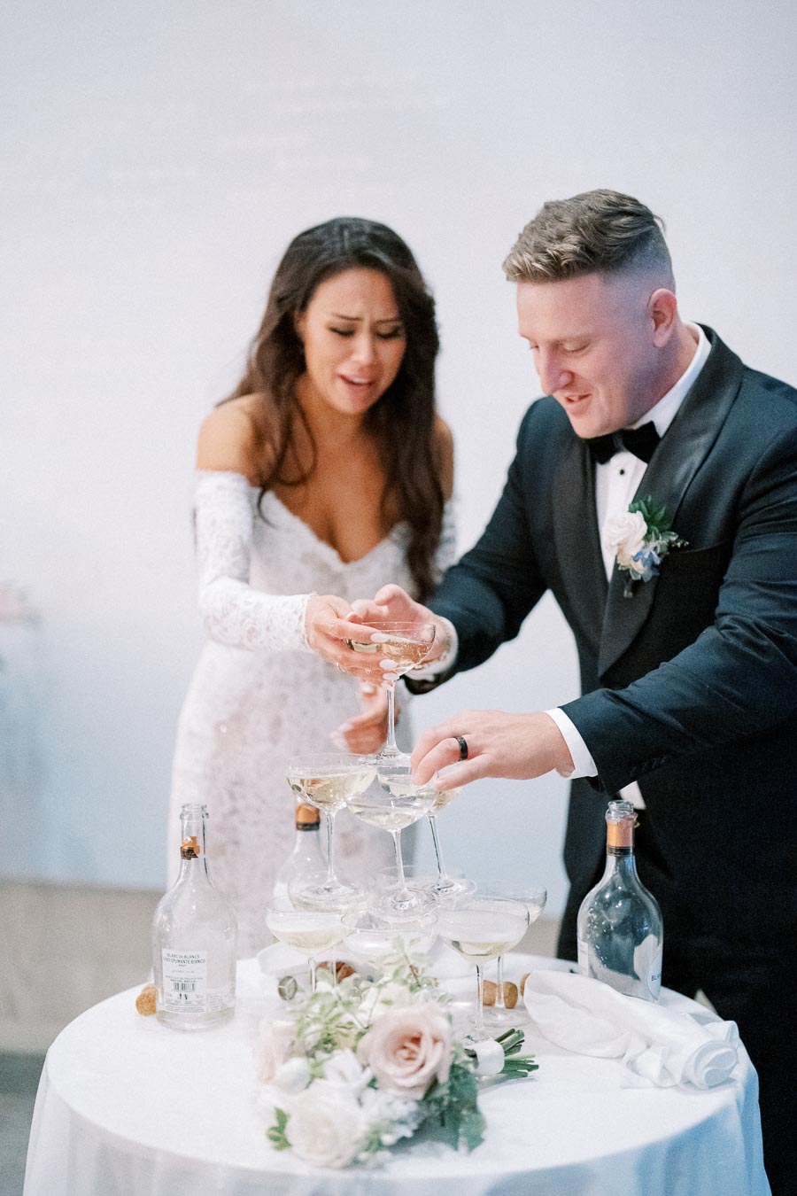 A bride and groom in formal wedding attire pour champagne into a tiered glass tower at their wedding reception, with a bouquet of white and blush pink flowers on the table.