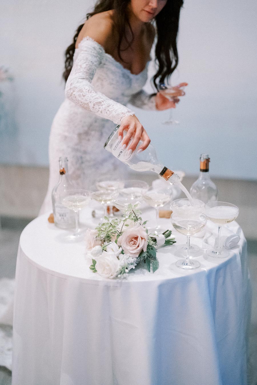 A bride in a lace wedding dress pours champagne into glasses on a round table decorated with pink and white roses, creating a romantic wedding setting.