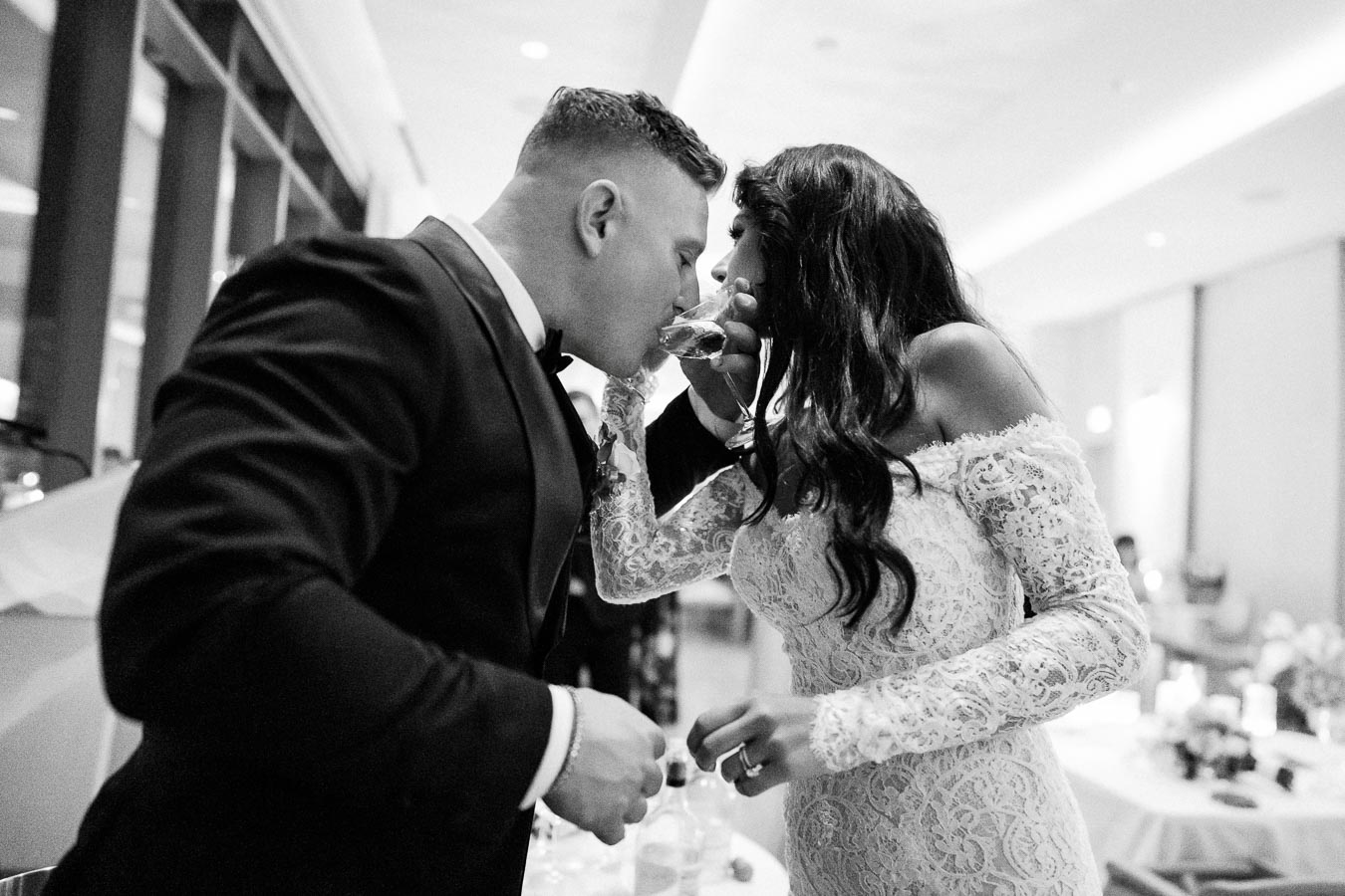 Elegant wedding couple sharing a toast, bride in a lace gown and groom in a black tuxedo, celebrating their special day in a beautifully decorated venue.