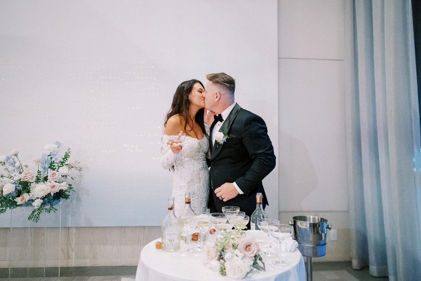 A newlywed couple shares a romantic kiss during their wedding celebration, standing beside a table adorned with champagne glasses and floral arrangements. The bride is in an elegant lace gown, and the groom is dressed in a classic black tuxedo.
