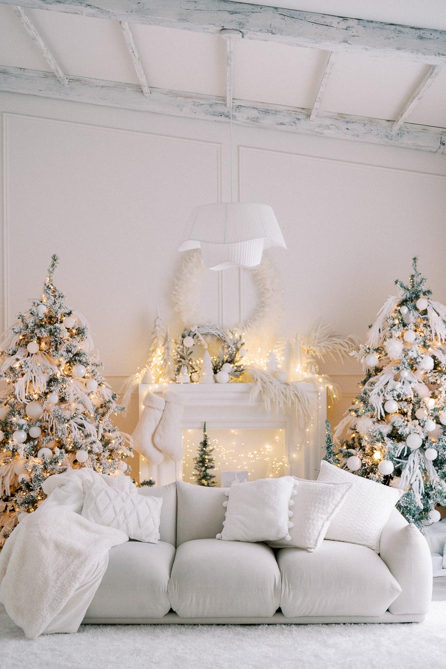 Elegant living room decorated for Christmas with white-themed decor. Features a cozy white sofa, two Christmas trees adorned with white ornaments and twinkling lights, and a mantel with festive stockings and a wreath. Bright, airy atmosphere.