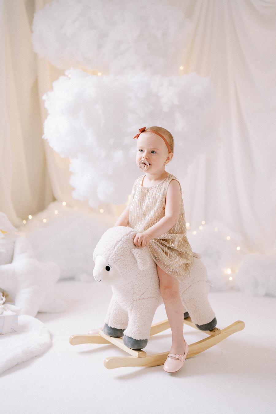 A toddler in a sparkling dress and headband sitting on a plush llama rocking toy in a dreamy, cloud-themed nursery with white drapes and soft lighting.
