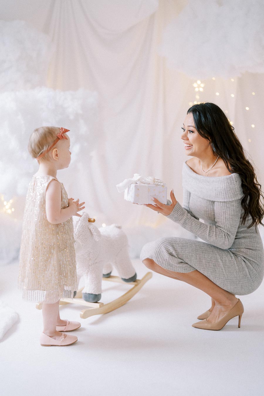 A woman in a gray dress kneels and smiles while giving a wrapped gift to a toddler in a sparkly dress and headband, in a white, festive setting with a plush rocking toy.