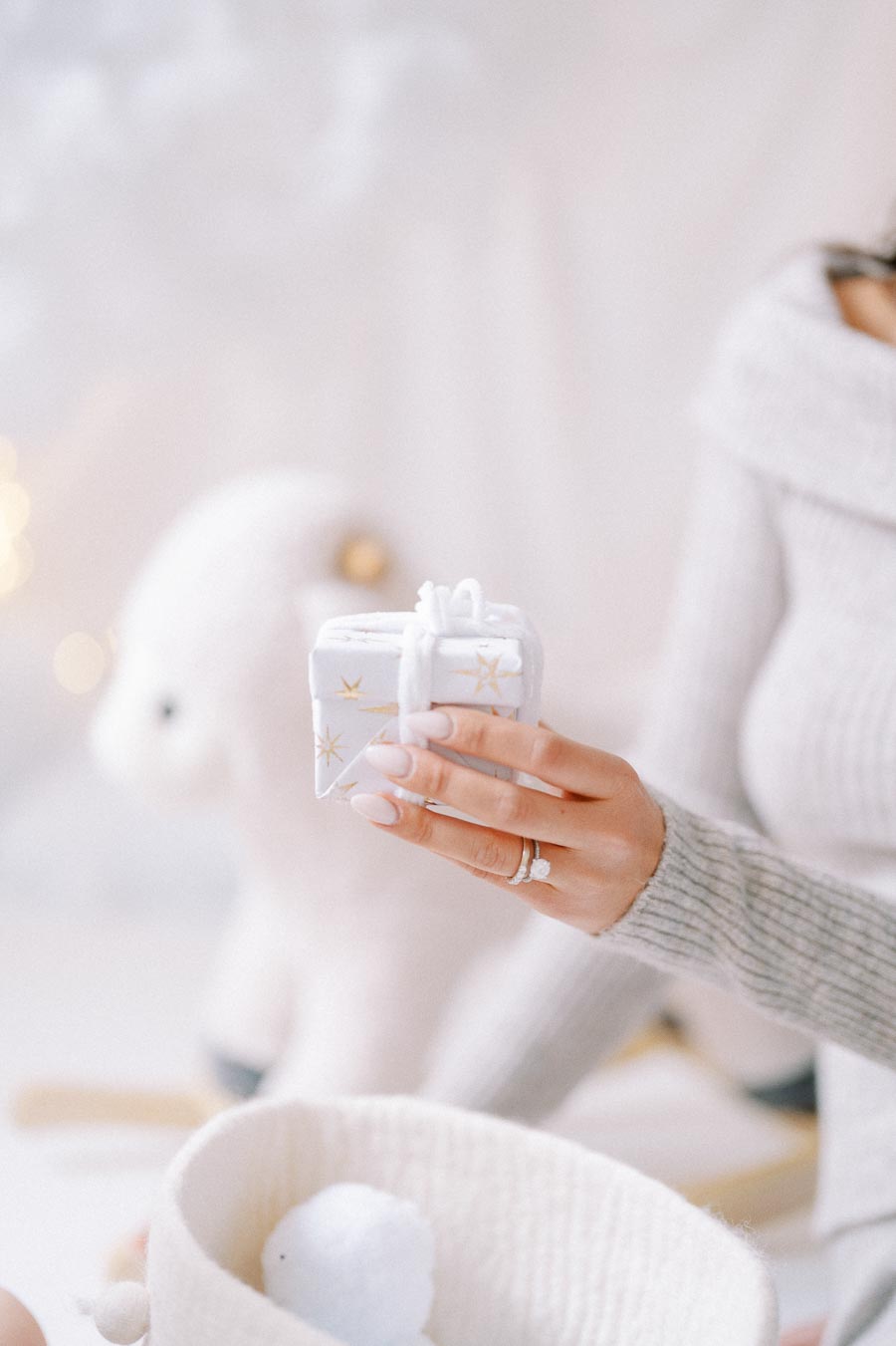 A person holding a small white gift box with gold star patterns, wearing a cozy sweater in a softly lit, festive setting with a teddy bear and basket in the background.