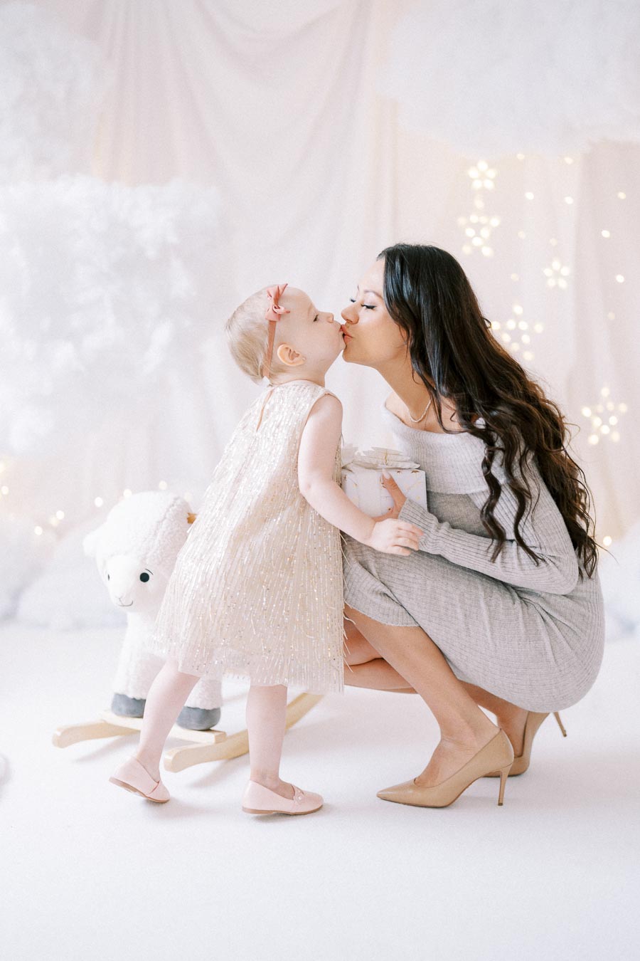 Mother and daughter sharing a loving kiss in a bright, festive setting with twinkling lights and a plush toy in the background.