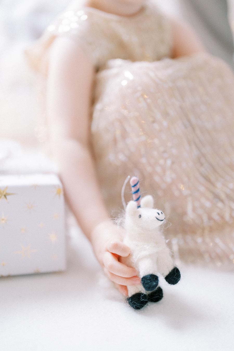 Child holding a small white toy unicorn with blue hooves, next to a gift box wrapped in star-patterned paper, wearing a sparkly gold dress.