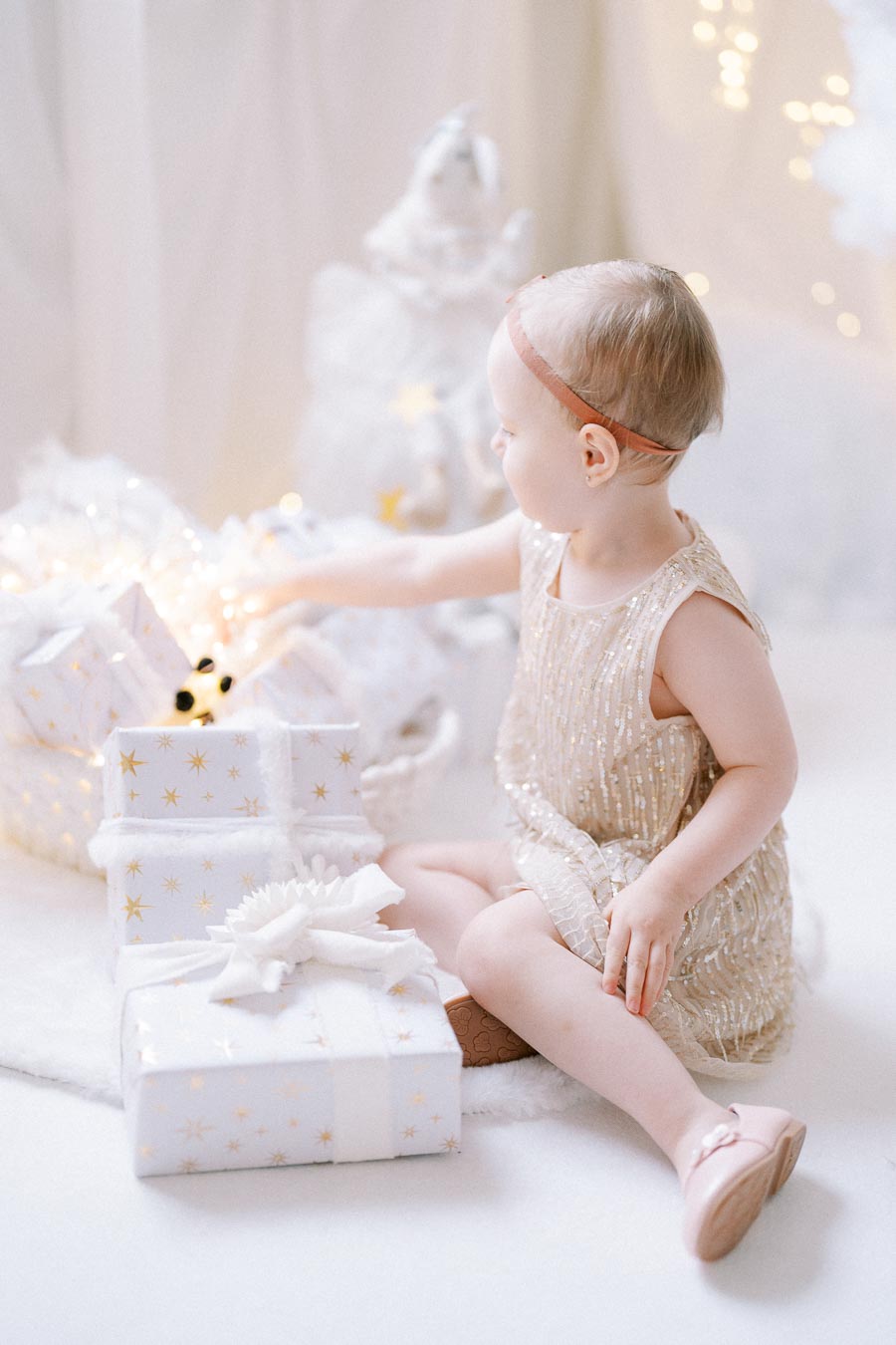 A toddler in a sparkly dress sits on the floor with wrapped Christmas presents, surrounded by soft, twinkling lights in a festive, cozy setting.