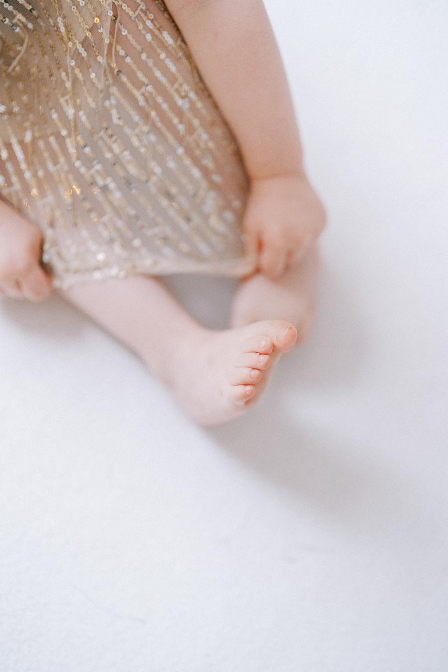 Close-up of a baby's legs and feet, wearing a sparkly, gold-sequined dress, on a white background.
