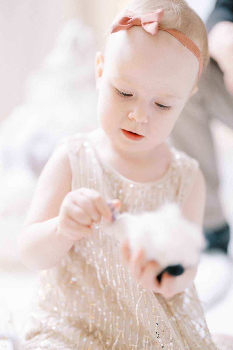 A young child wearing a sparkly dress and pink headband, focused on playing with a fluffy toy, in a softly lit room.