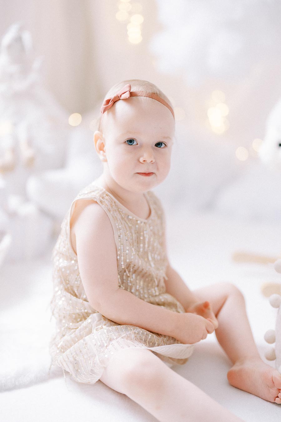 A baby in a sparkly dress with a pink bow headband sitting on a soft white surface, with blurred festive lights in the background.