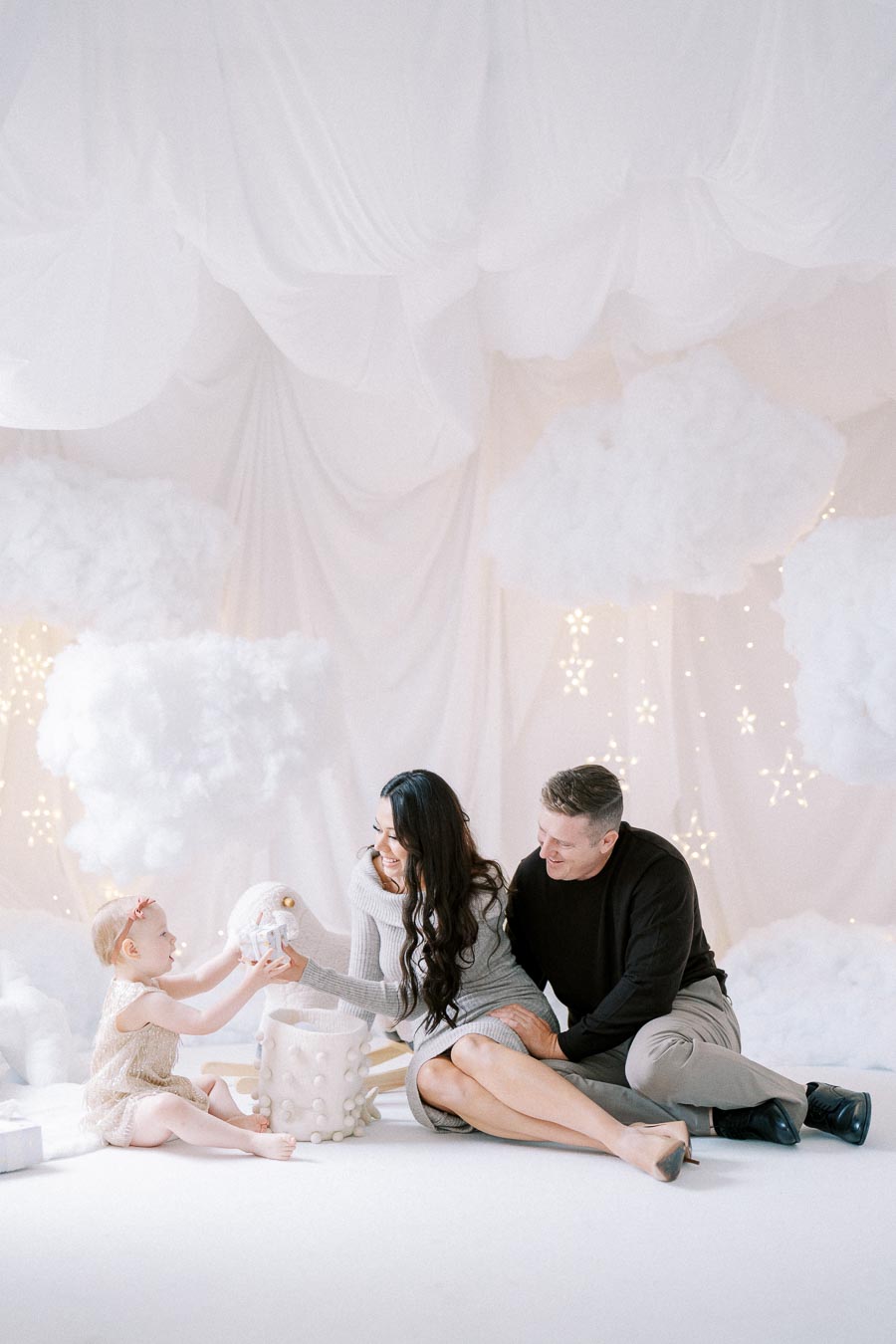 A family enjoying a whimsical, cloud-themed photoshoot in a soft, pastel setting with starry lights, featuring two adults and a child exchanging gifts.