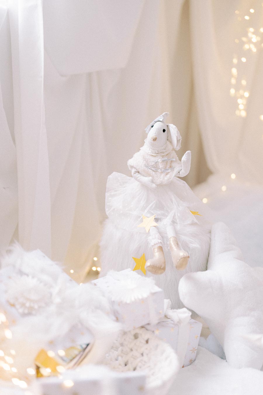 Plush mouse toy in elegant white dress with star accents sitting on white furry surface, surrounded by twinkling fairy lights and wrapped gifts in a cozy, dreamy setting.