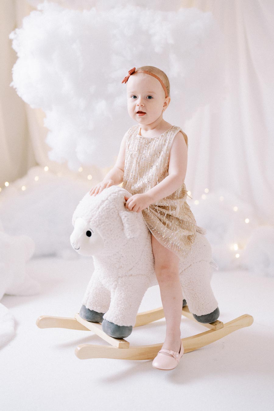 Toddler wearing a gold dress and headband riding a plush rocking sheep in a dreamy, light-filled nursery setting with soft cloud decorations.