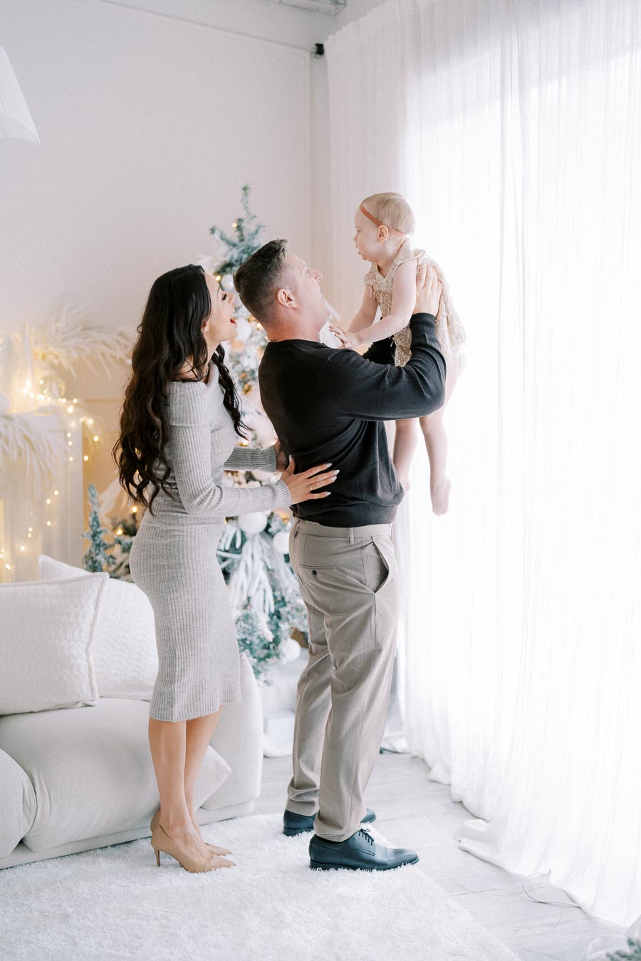 Happy family in a modern living room with Christmas decorations, parents playing with their baby near a large window.