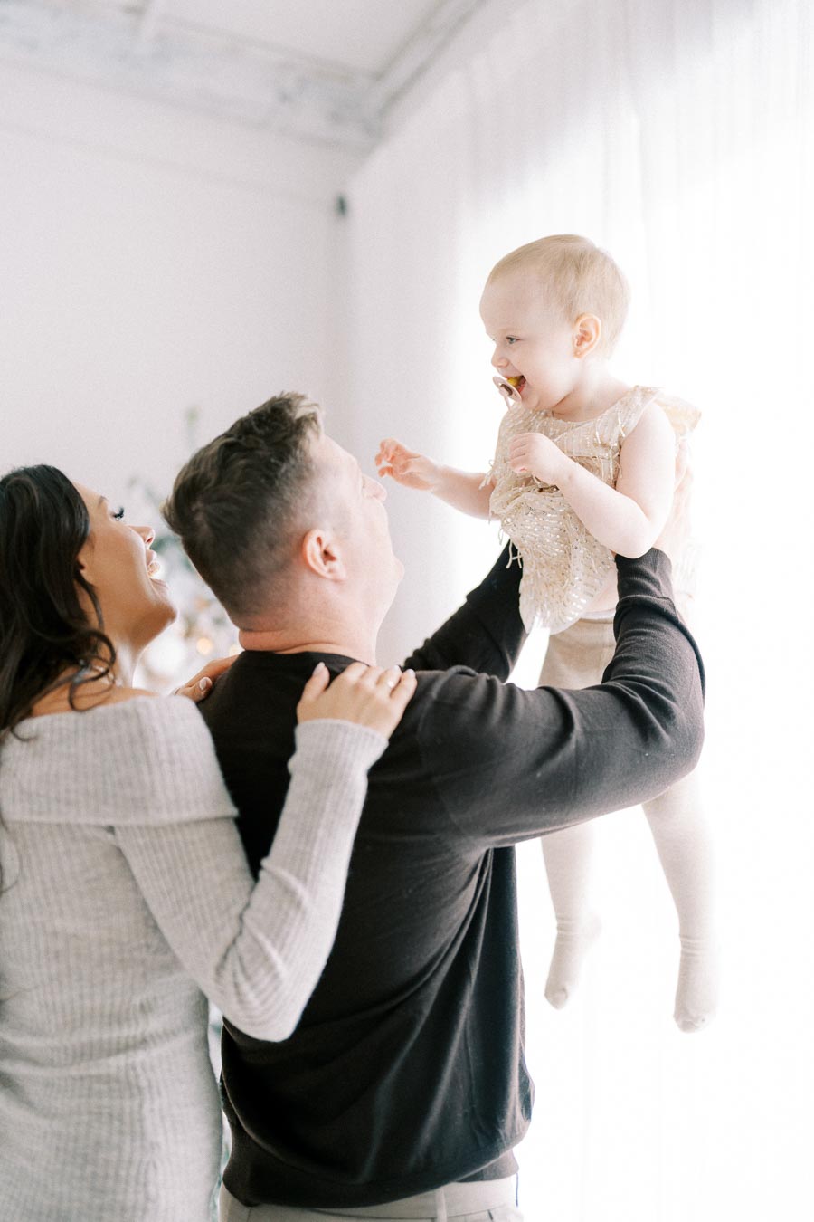 A joyful family moment with a father lifting his baby daughter as the smiling mother watches, set in a bright and airy room.