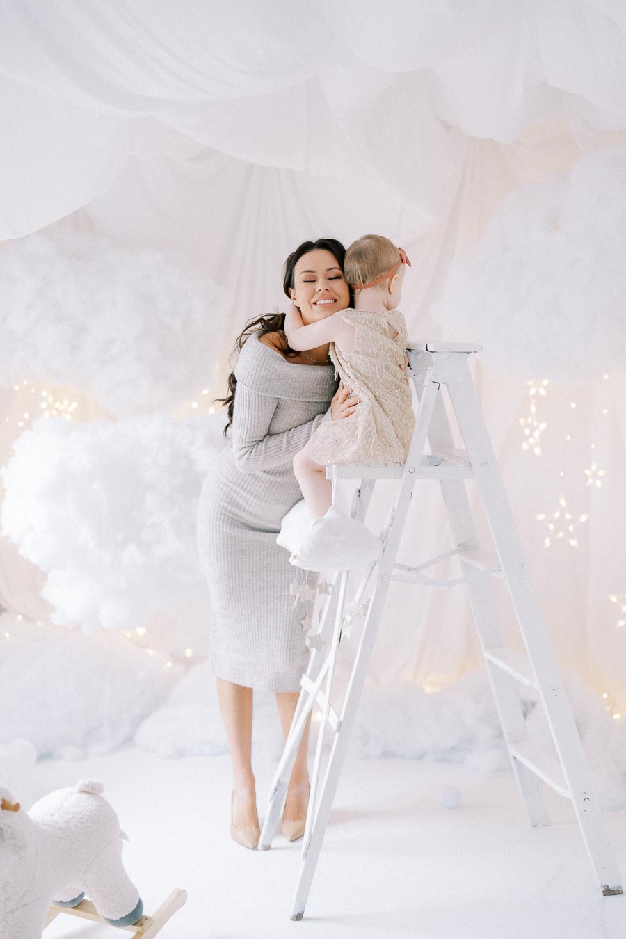 A woman smiling while hugging a child on a ladder in a whimsical, snowy-themed room with cotton clouds and twinkling lights.
