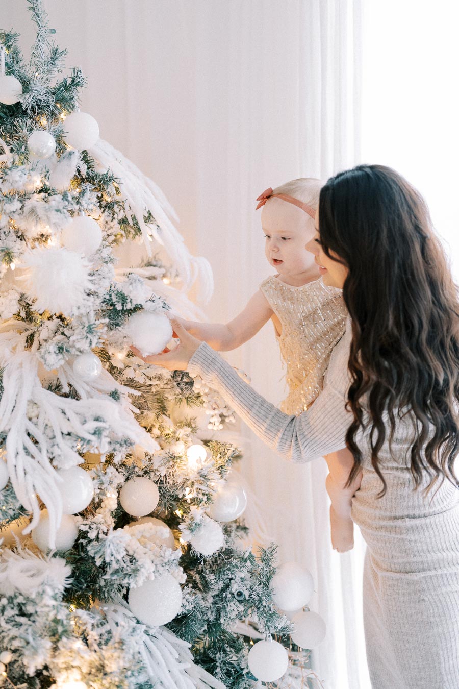 A woman and child decorating a snowy Christmas tree with white ornaments, glowing holiday lights, and festive decorations in a cozy indoor setting.