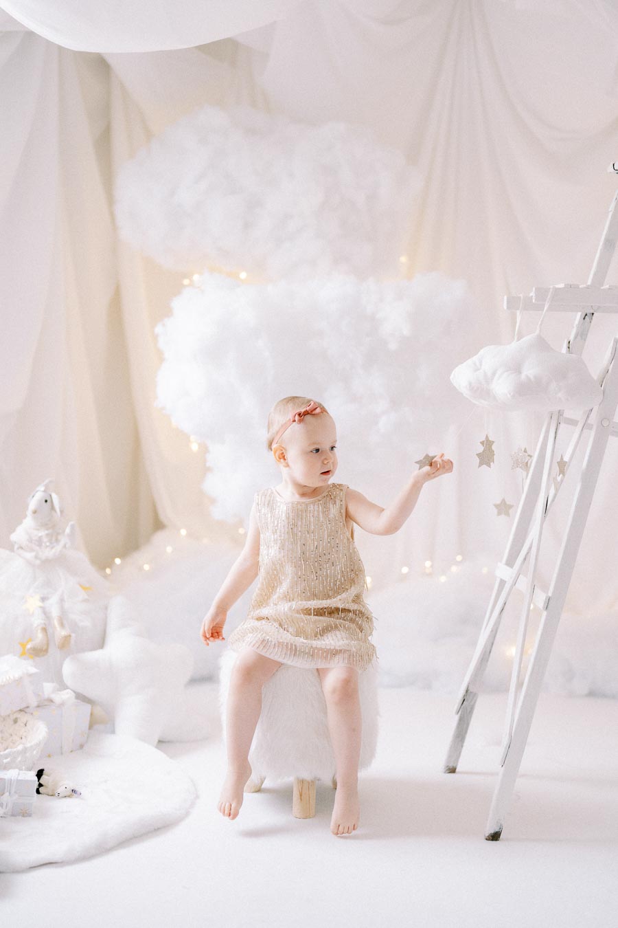 A toddler wearing a glittery dress sits in a whimsical cloud-themed setting, surrounded by soft, white, fluffy decorations and a white ladder adorned with hanging stars, creating a dreamy atmosphere.