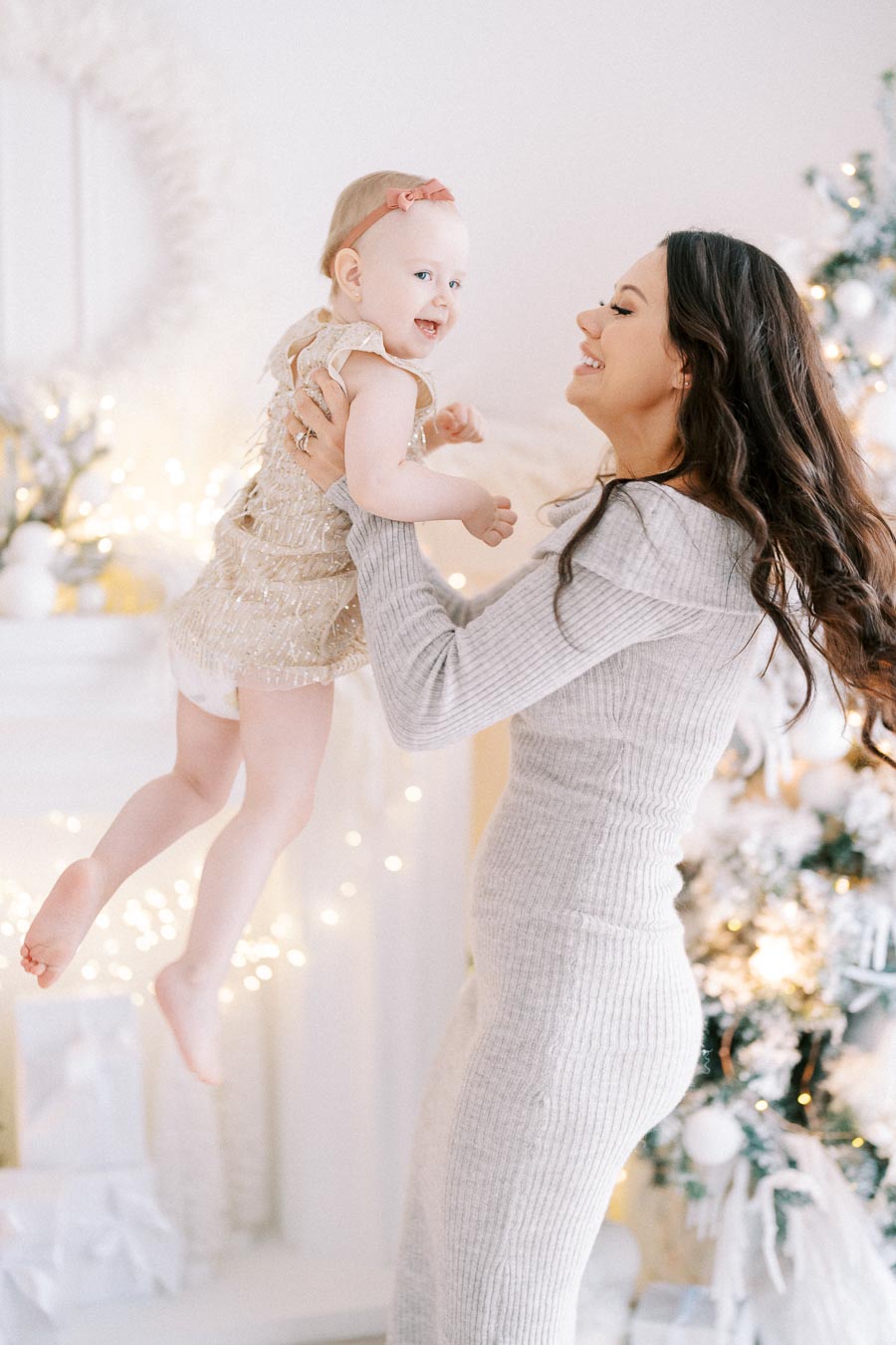 A joyful woman lifting a smiling baby in a festive, decorated room with Christmas lights and a tree in the background.