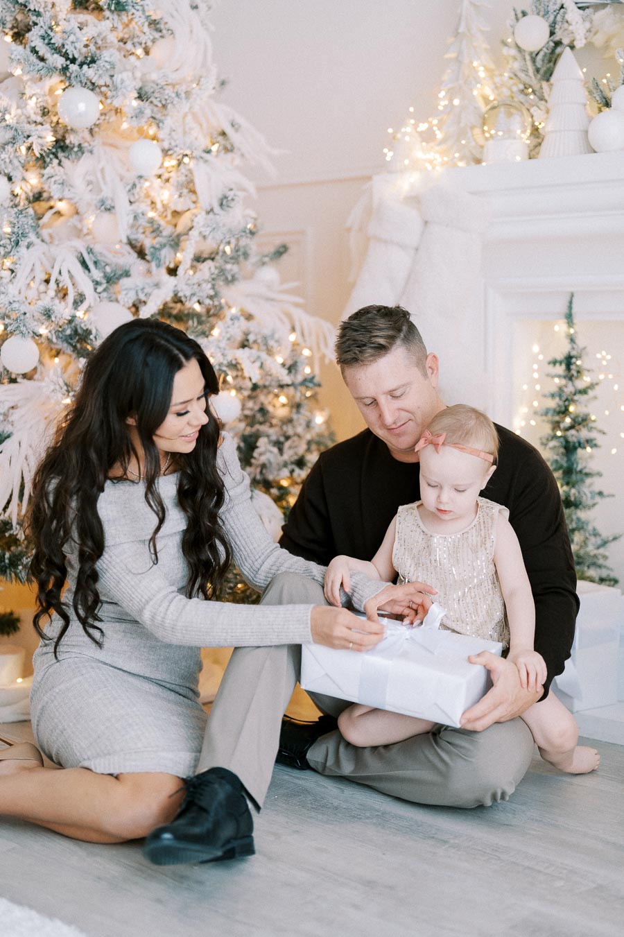 Family sitting together opening a Christmas gift in a beautifully decorated living room, featuring a snow-covered Christmas tree and festive lights.