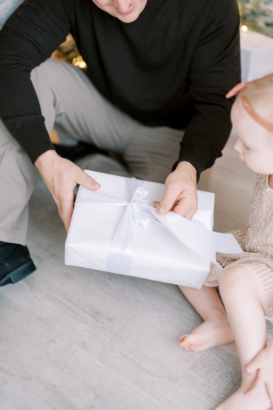A person in a black sweater hands a white gift box with a satin ribbon to a seated child in a festive setting, implying a joyful, holiday atmosphere.