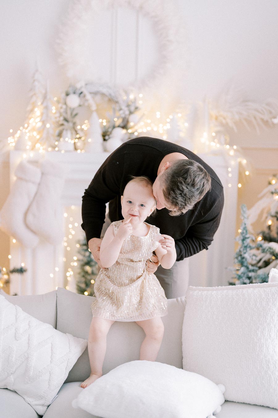 Father holding baby girl wearing a sparkly dress in a festive, decorated living room with white and gold Christmas ornaments.