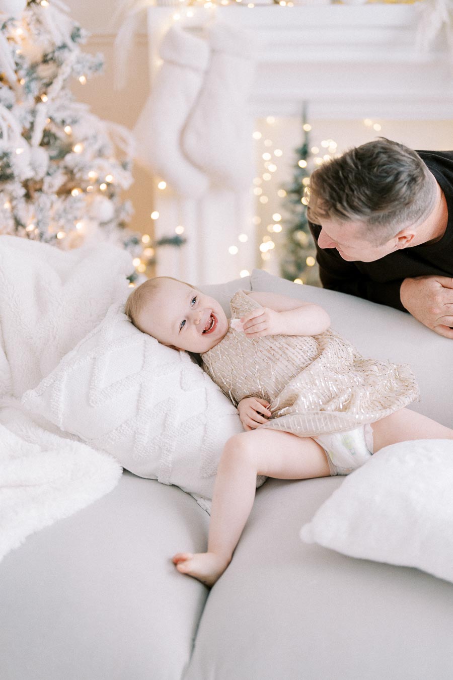 A joyful toddler in a festive gold dress laughing on a cozy couch with a Christmas tree in the background and soft white pillows, creating a warm holiday atmosphere.