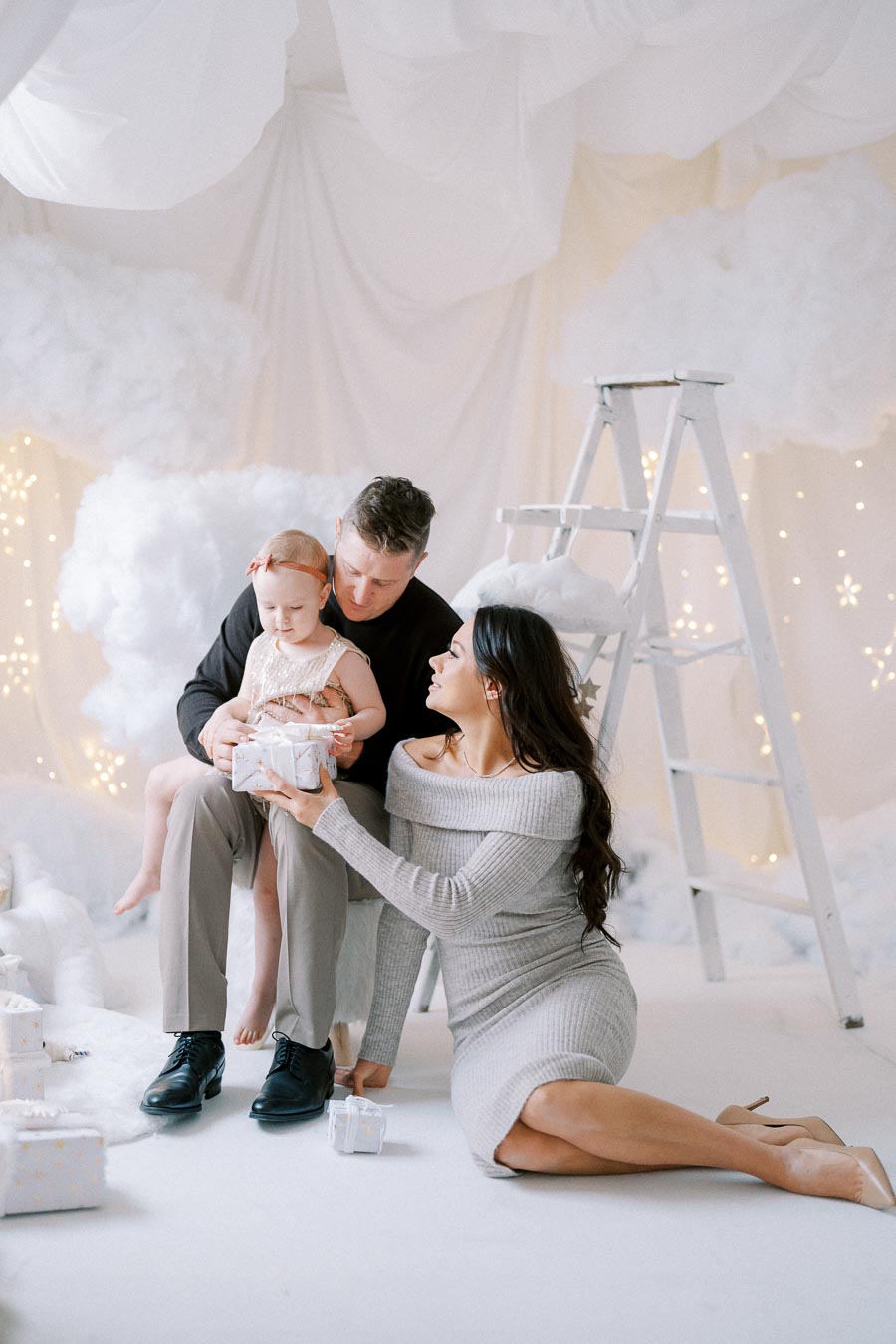 Family celebrating in winter wonderland setting with child holding a gift, surrounded by white decorations and fairy lights.