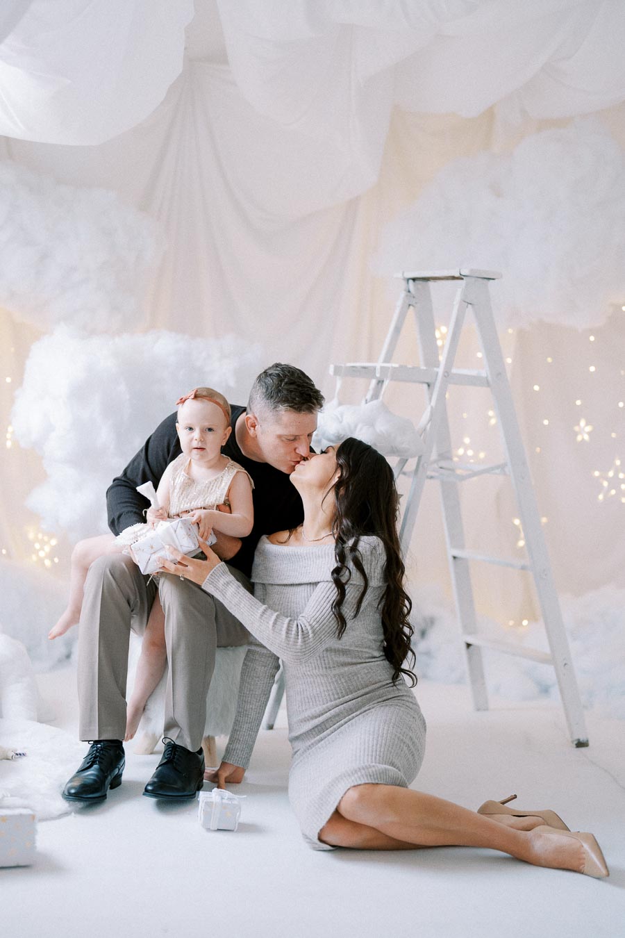 A happy family celebrating in a winter wonderland theme, with a father holding a child and sharing a kiss with the mother, surrounded by white clouds and twinkling lights.