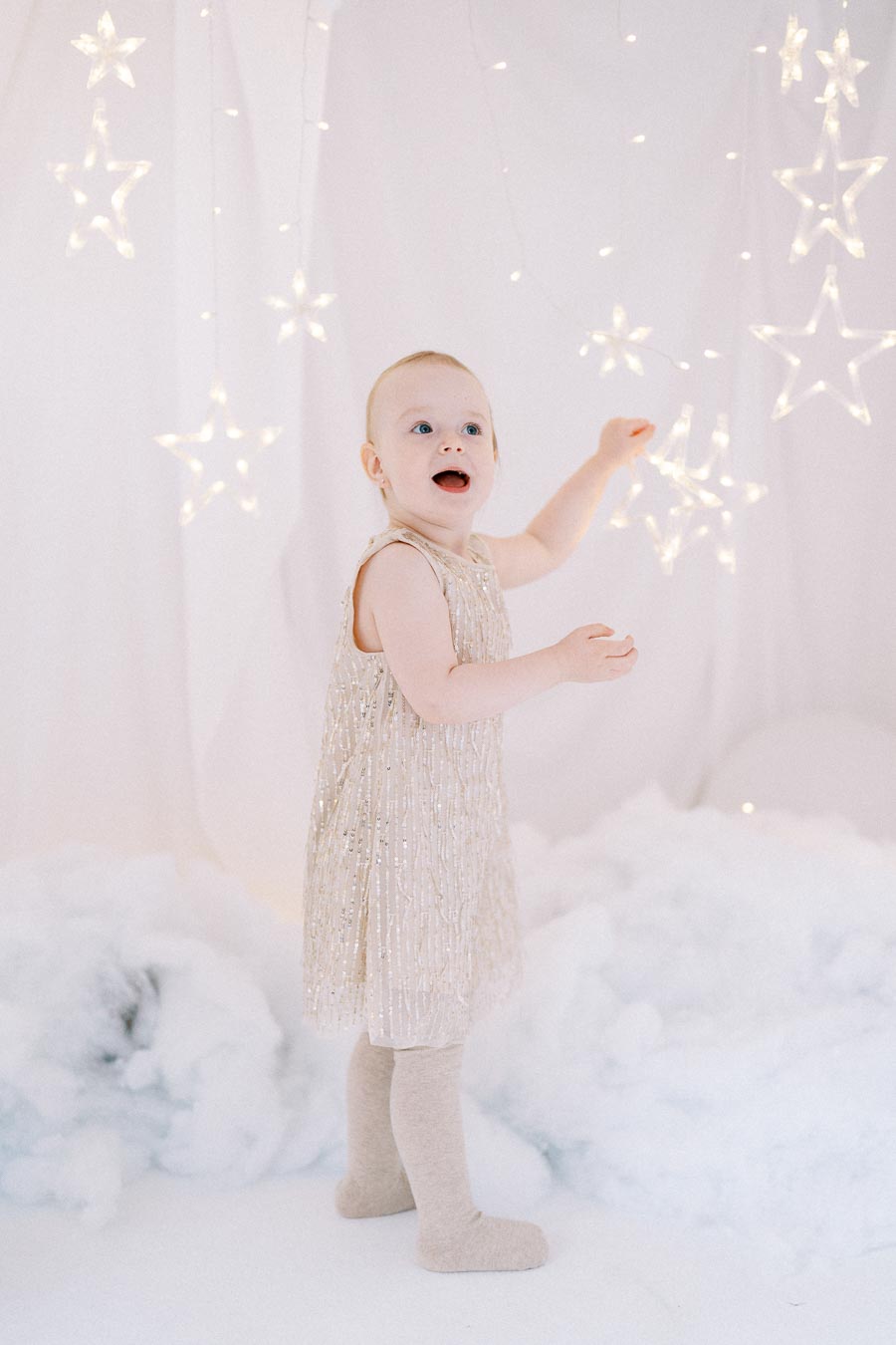 A young child in a sparkly dress stands surrounded by glowing star lights and fluffy clouds, creating a dreamy and magical atmosphere.