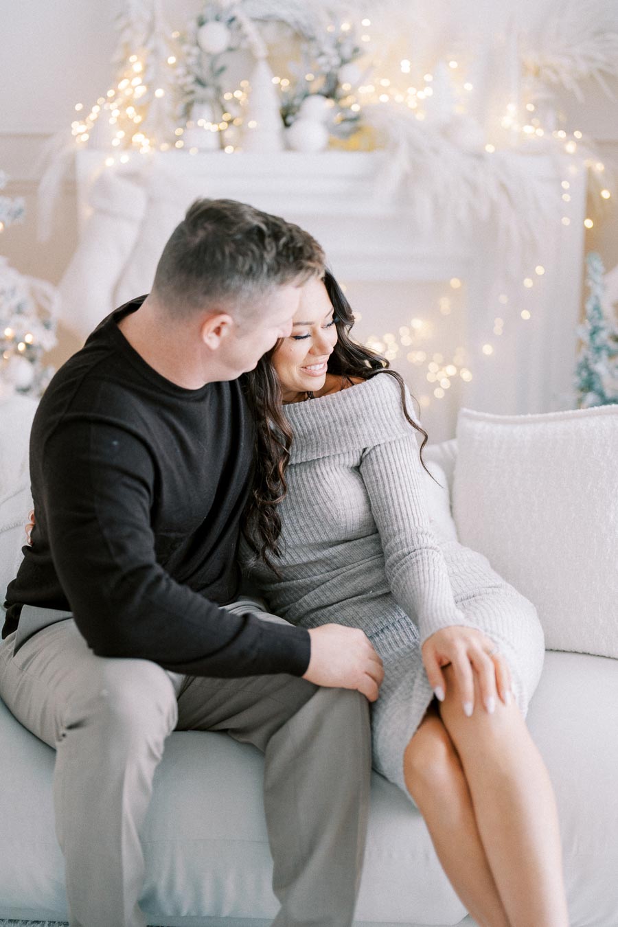 A couple sitting closely on a cozy white sofa, smiling and enjoying a festive holiday setting with twinkling lights and elegant winter decor in the background.