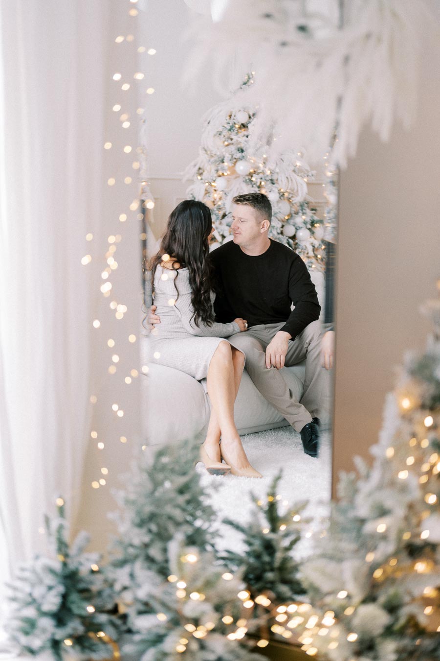 A couple sitting on a couch, surrounded by Christmas decorations and festive lights, with a decorated tree in the background.