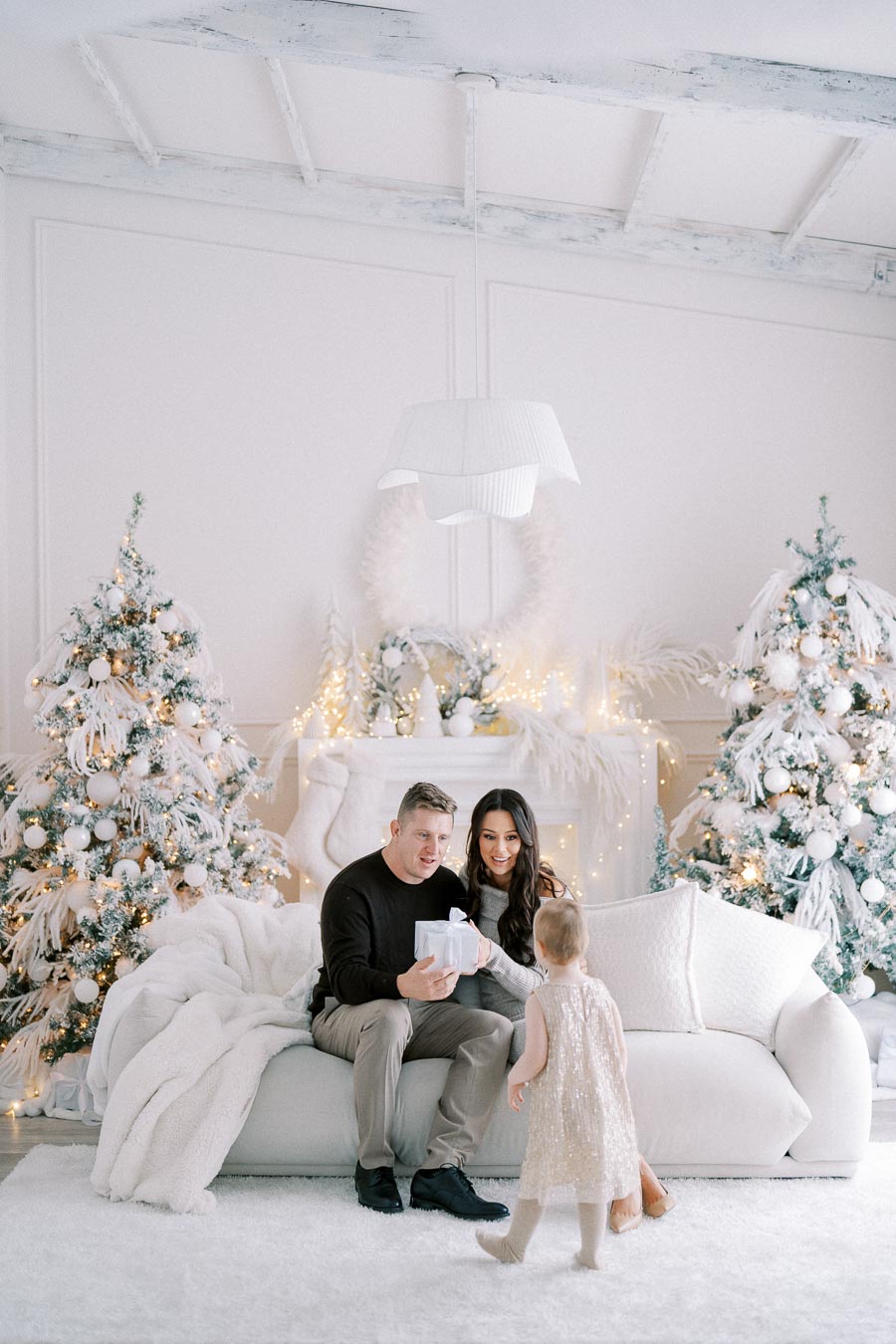 A family enjoying a festive moment in a beautifully decorated living room with Christmas trees and lights. A young child approaches parents who are seated on a white sofa, holding a wrapped gift, creating a warm holiday atmosphere.