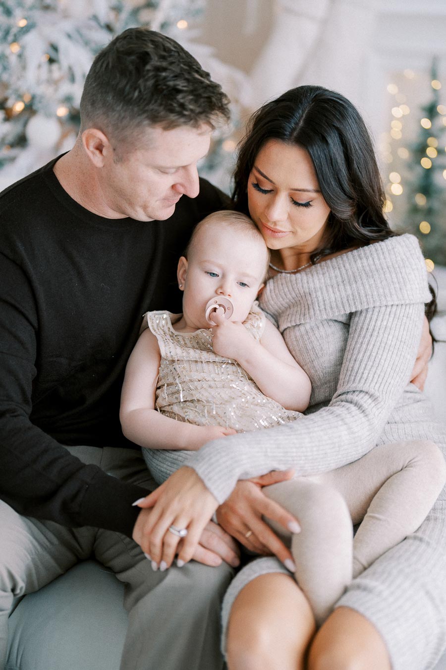 A loving family sitting together on a couch, with a father, mother, and young child dressed in cozy winter attire, in front of a softly lit, festive Christmas tree.
